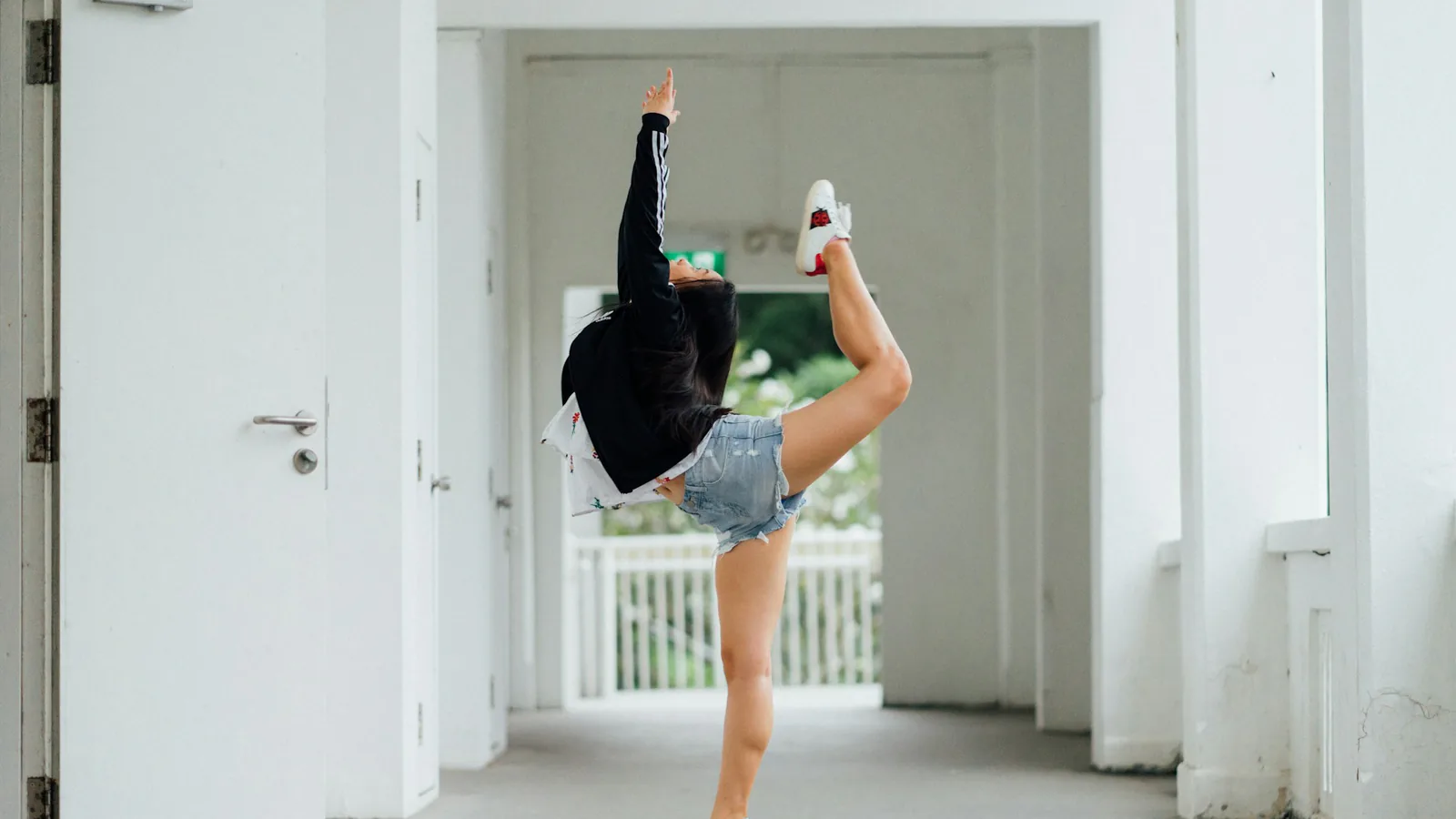 Eine Person in Jeansshorts und weißen Turnschuhen hebt ein angewinkeltes Bein und einen Arm in einem hellen Flur.