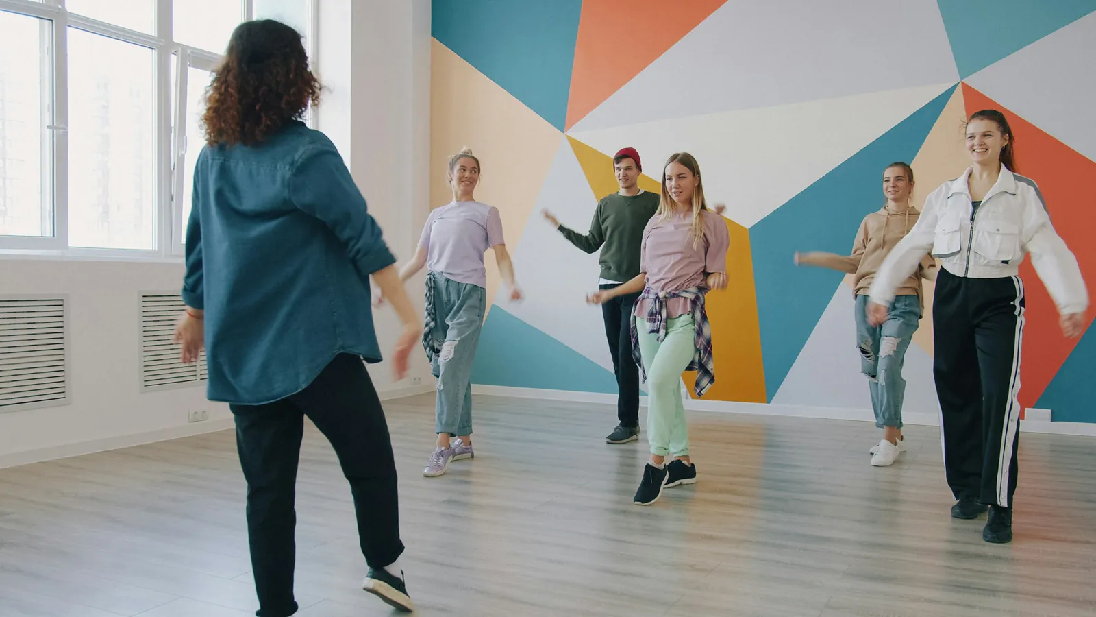 Eine Person steht einer tanzenden Gruppe in einem hellen Raum mit bunt geometrisch bemalter Wand gegenüber.