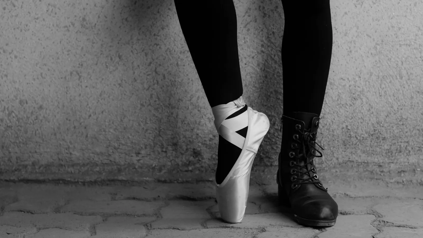 Nahaufnahme zweier Füße: ein heller Spitzenschuh und ein schwarzer Schnürstiefel auf Pflastersteinen.