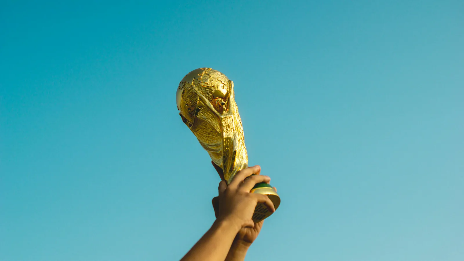 Hände halten einen goldfarbenen Pokal in Form des Fußball-WM-Pokals vor einem blauen Himmel hoch.
