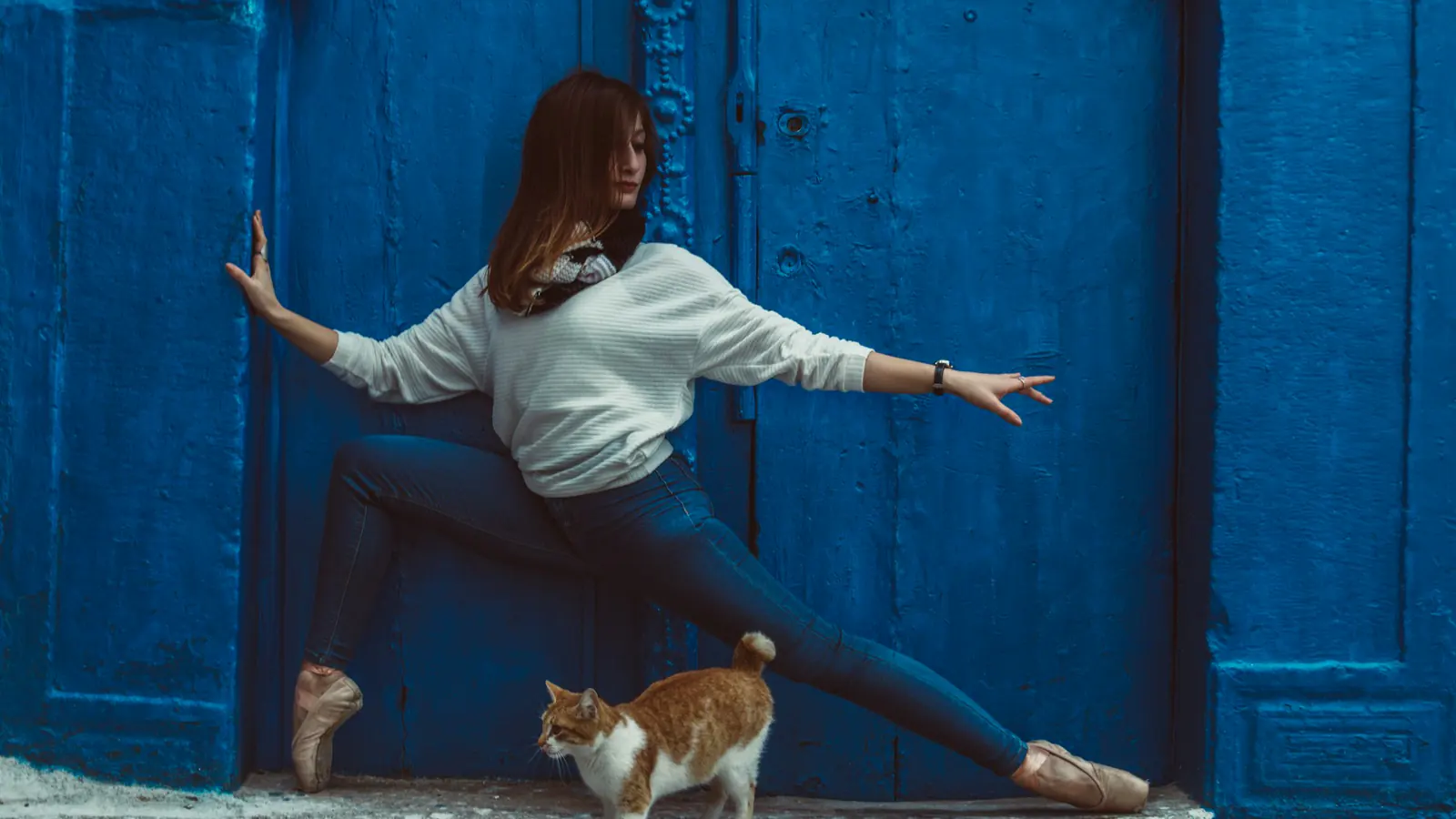 Eine Person posiert in Jeans und Spitzenschuhen vor einer blauen Tür; eine rot-weiße Katze läuft vor ihren Beinen.
