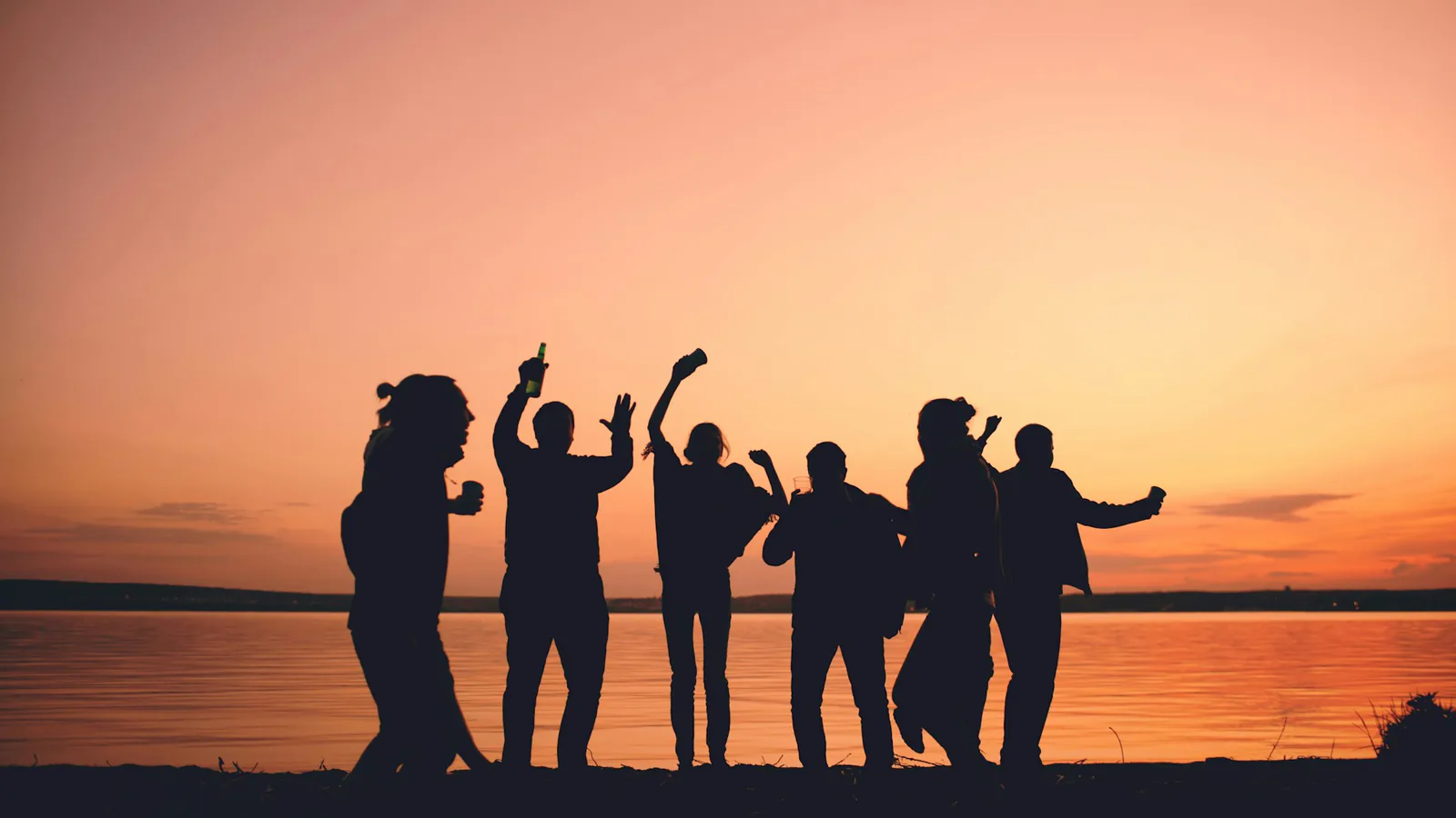Silhouetten mehrerer Menschen mit erhobenen Armen am Wasser vor einem orangefarbenen Abendhimmel.