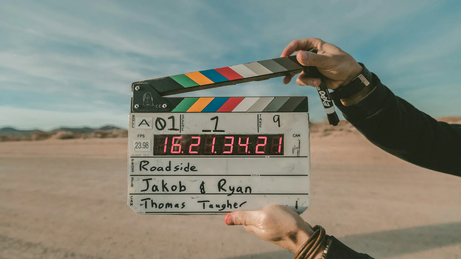 Zwei Hände halten eine geöffnete Filmklappe mit farbigen Streifen und digitaler Anzeige vor einer sandigen Landschaft.