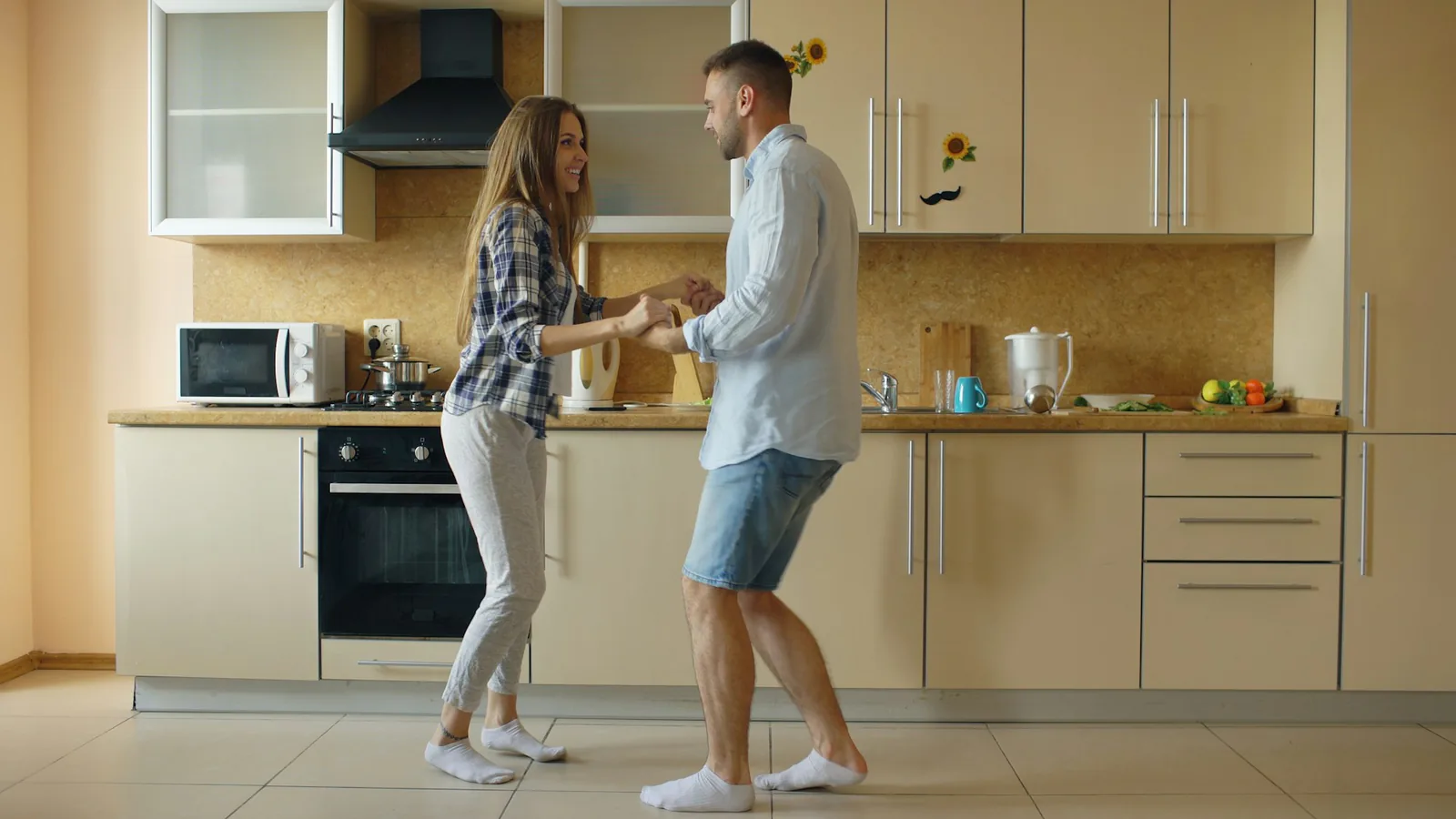 Zwei Menschen tanzen in einer Küche, halten sich an den Händen und tragen Socken auf dem Fliesenboden.