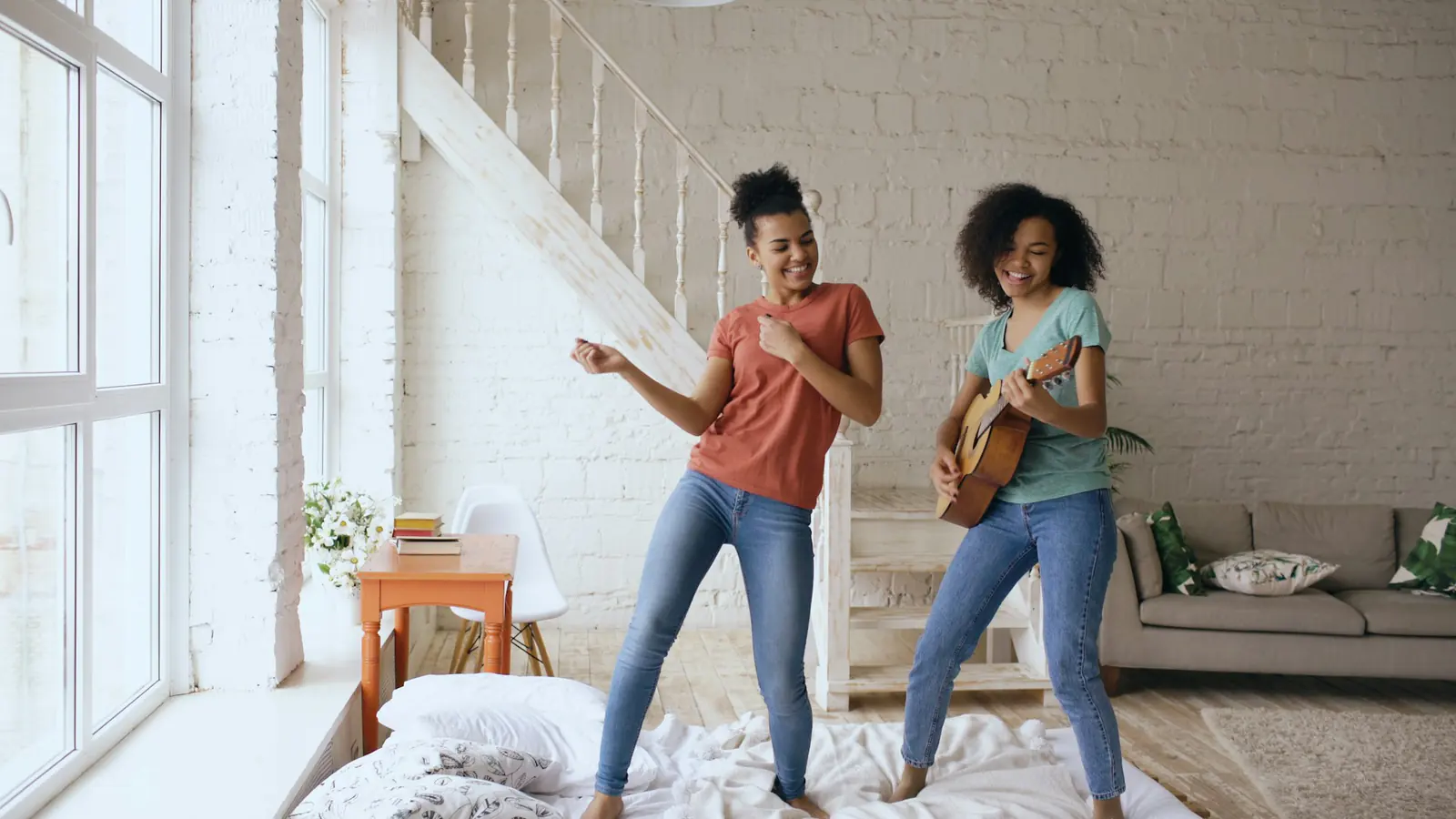 Zwei Menschen stehen barfuß auf einem Bett; eine Person hält eine Ukulele, die andere bewegt ihre angewinkelten Arme.