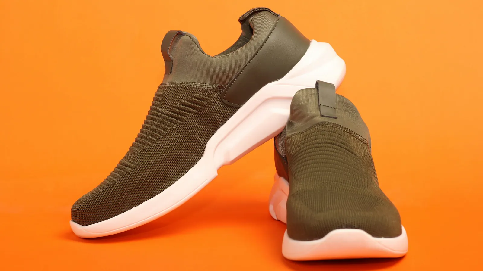 Zwei olivgrüne Schlupfschuhe mit weißen Sohlen auf orangefarbenem Hintergrund, einer schräg auf den anderen gelehnt.