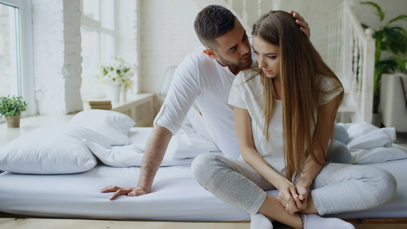 Zwei Menschen sitzen auf einem Bett; die hintere Person legt eine Hand an den Kopf der vorderen Person.